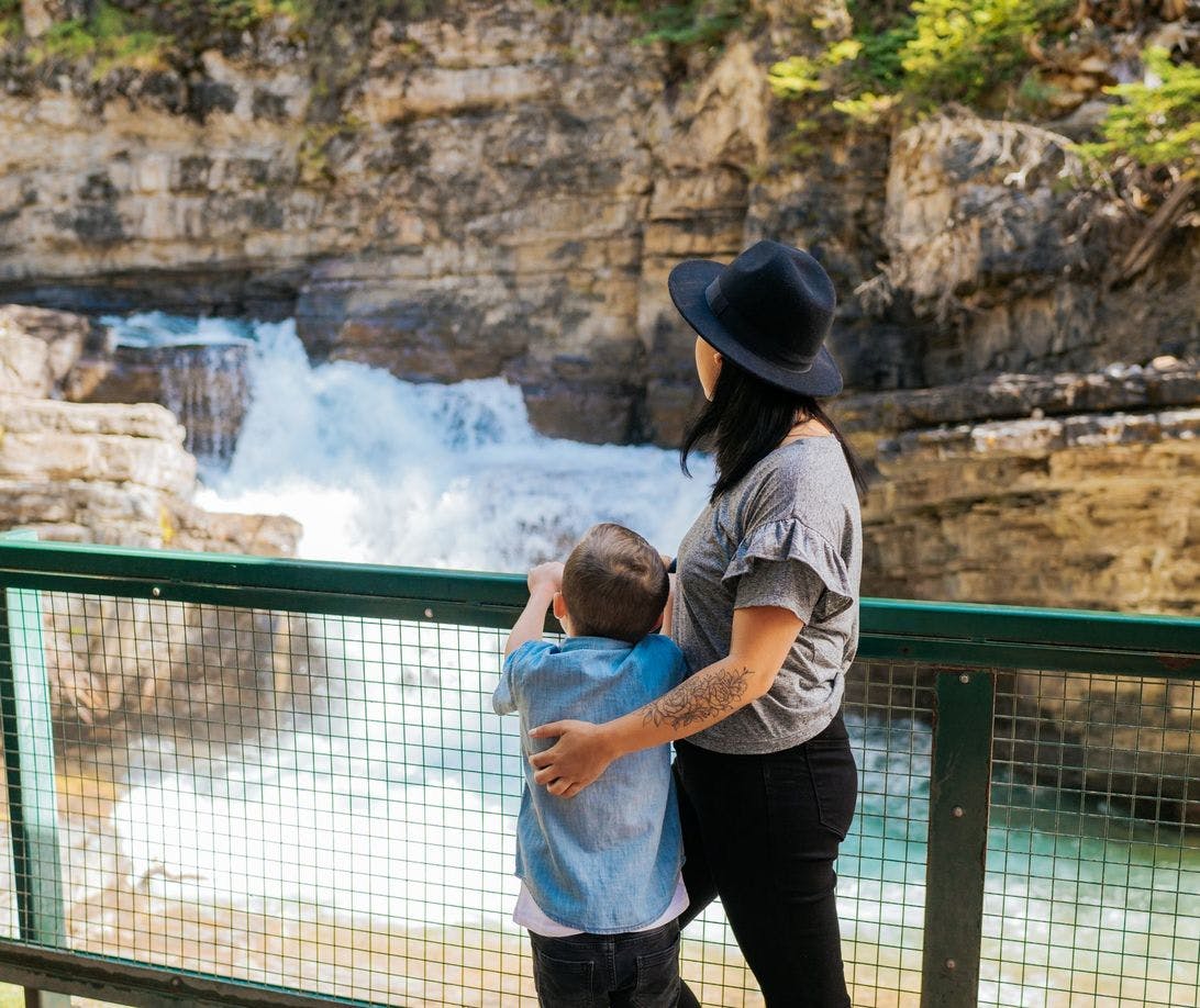 A mother and son stand alongside a railing overlooking a waterfall embedded in a large canyon
