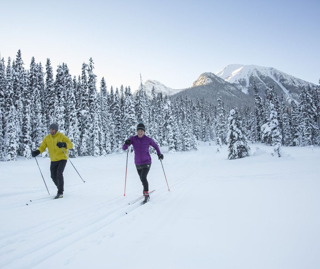 A couple cross-country skiing through fresh snow with mountains behind them