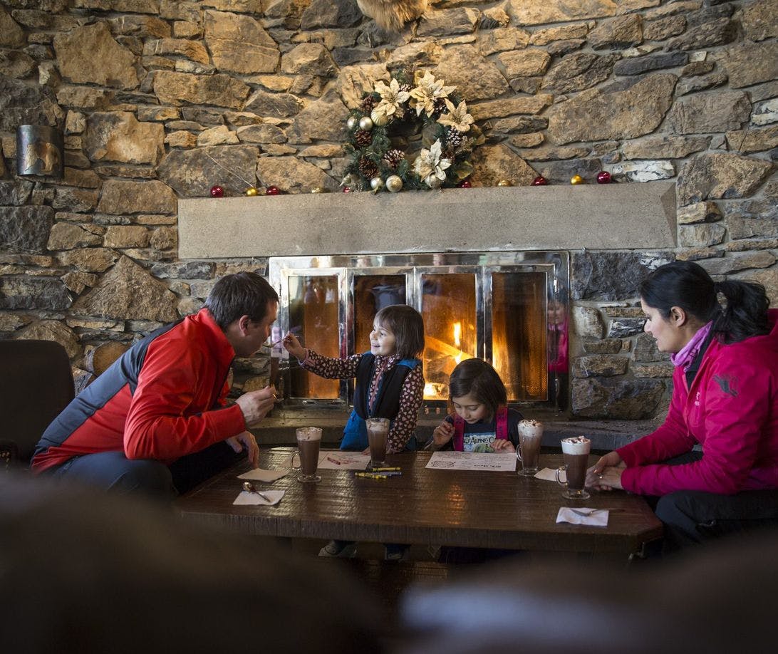A family of four take a break from skiing to enjoy hot chocolates in front of a crackling fire indoors