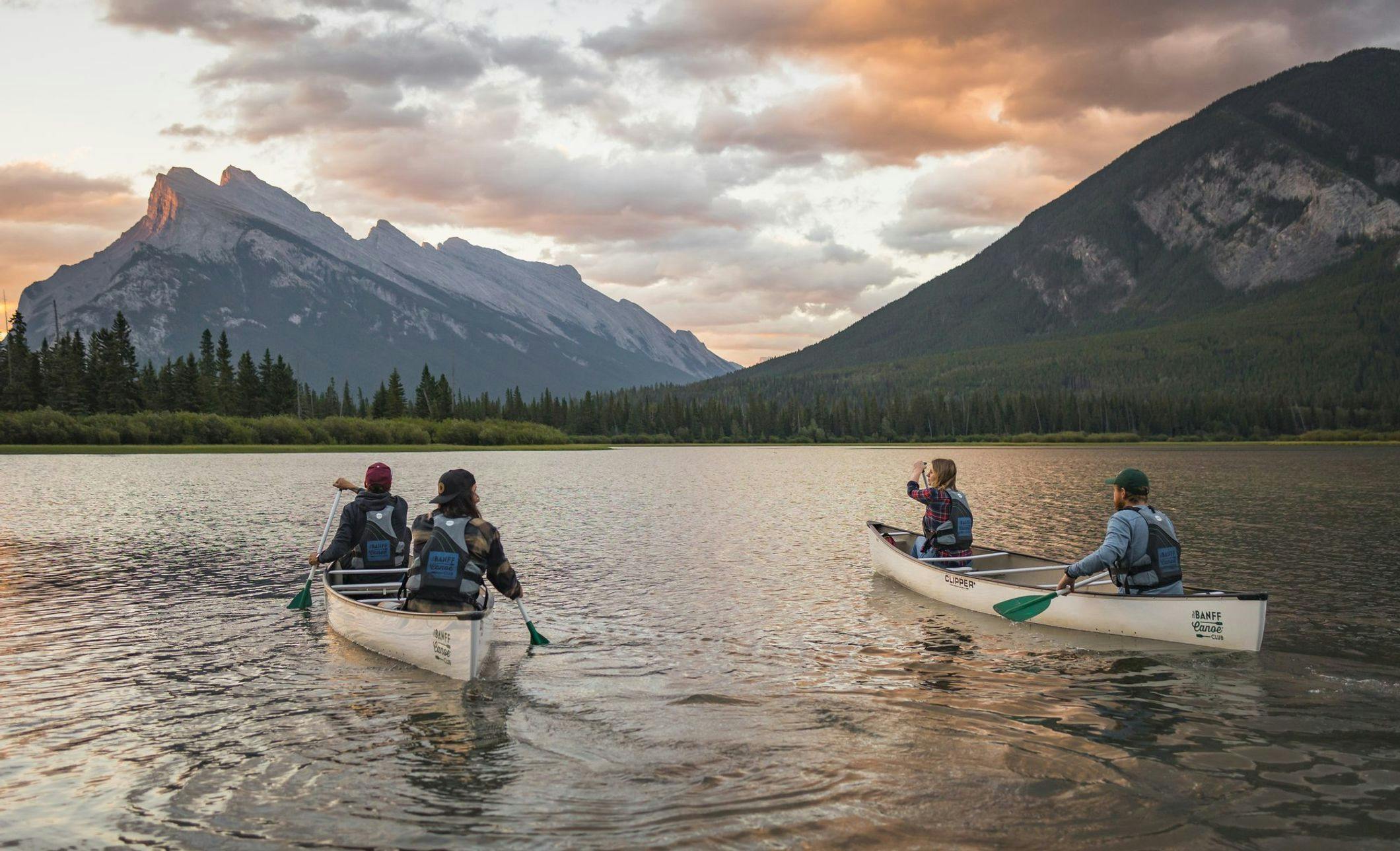 4 friends paddle in 2 canoes in an open lake surrounded by mountains during sunrise