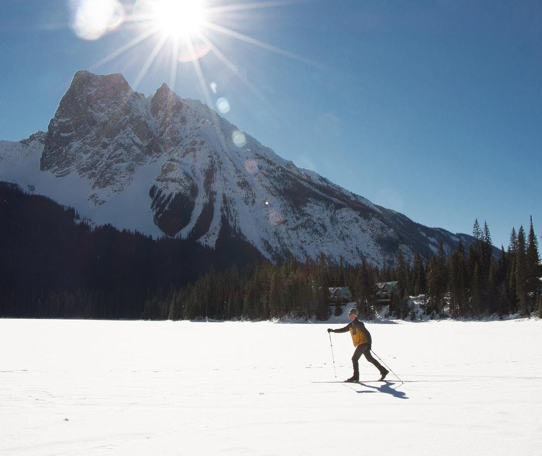 A man in a yellow jacket cross-country skis across a snow covered Emerald Lake with mountains and sunshine in the background
