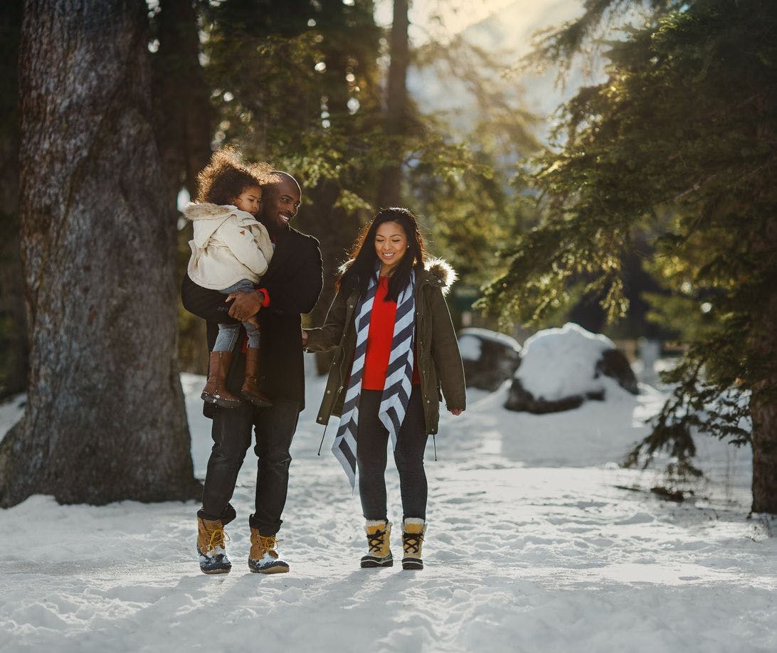 A family of 3 walking through the trees in the winter wearing boots, coats, and scarves