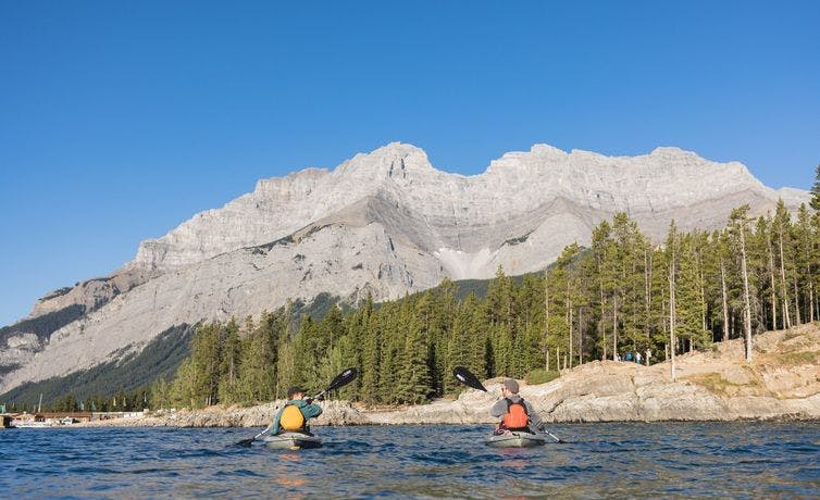 Two young males paddle in kayaks on a large blue lake with mountains and trees surrounding