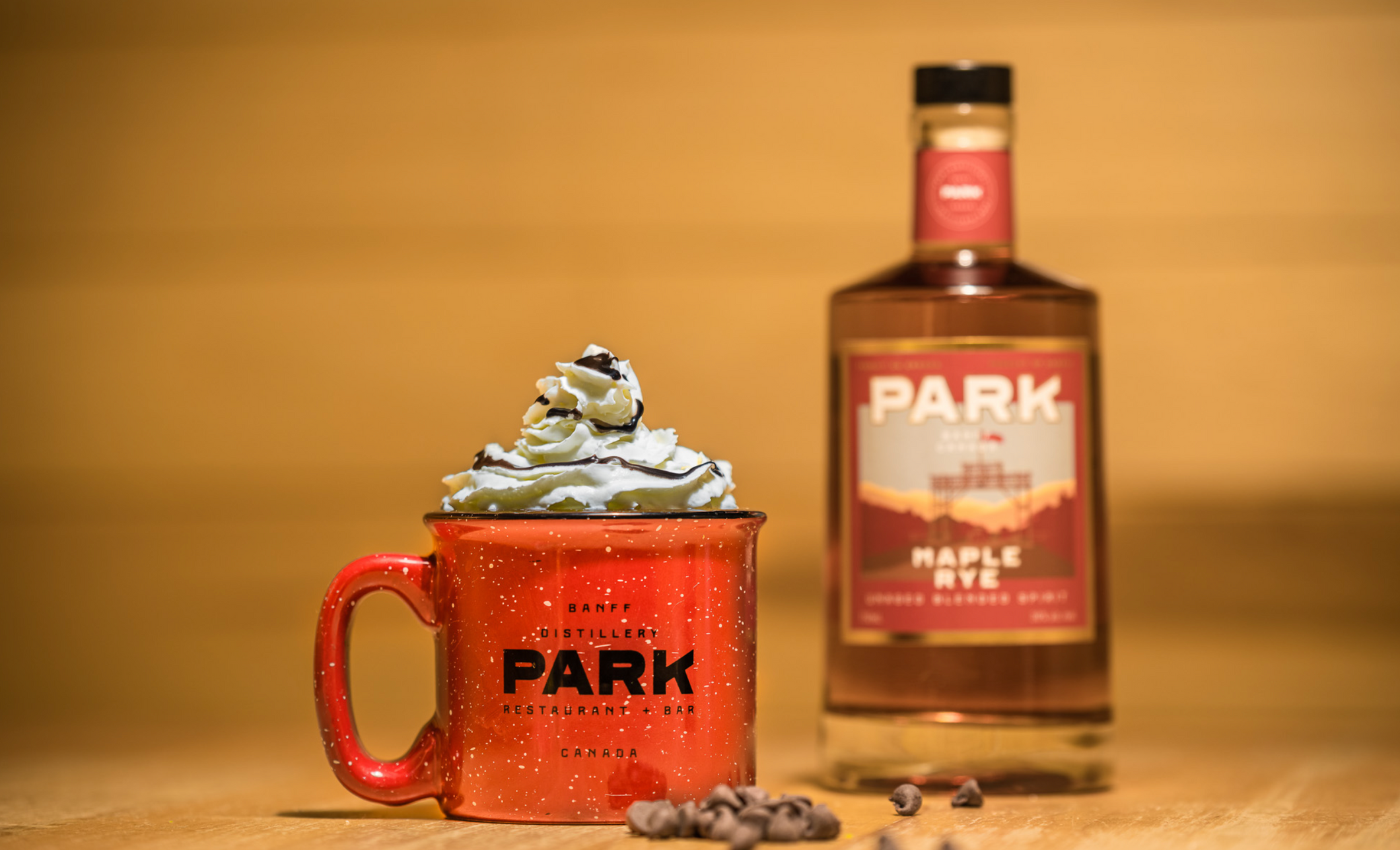 A hot chocolate in a red mug with whipped cream on a wooden table beside a liquor bottle.