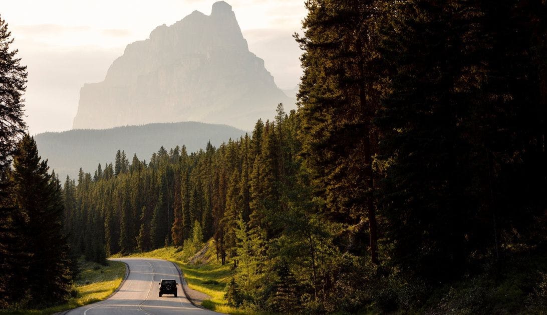 A Jeep drives down the Bow Valley Parkway in Banff National Park with Castle Mountain in the background.