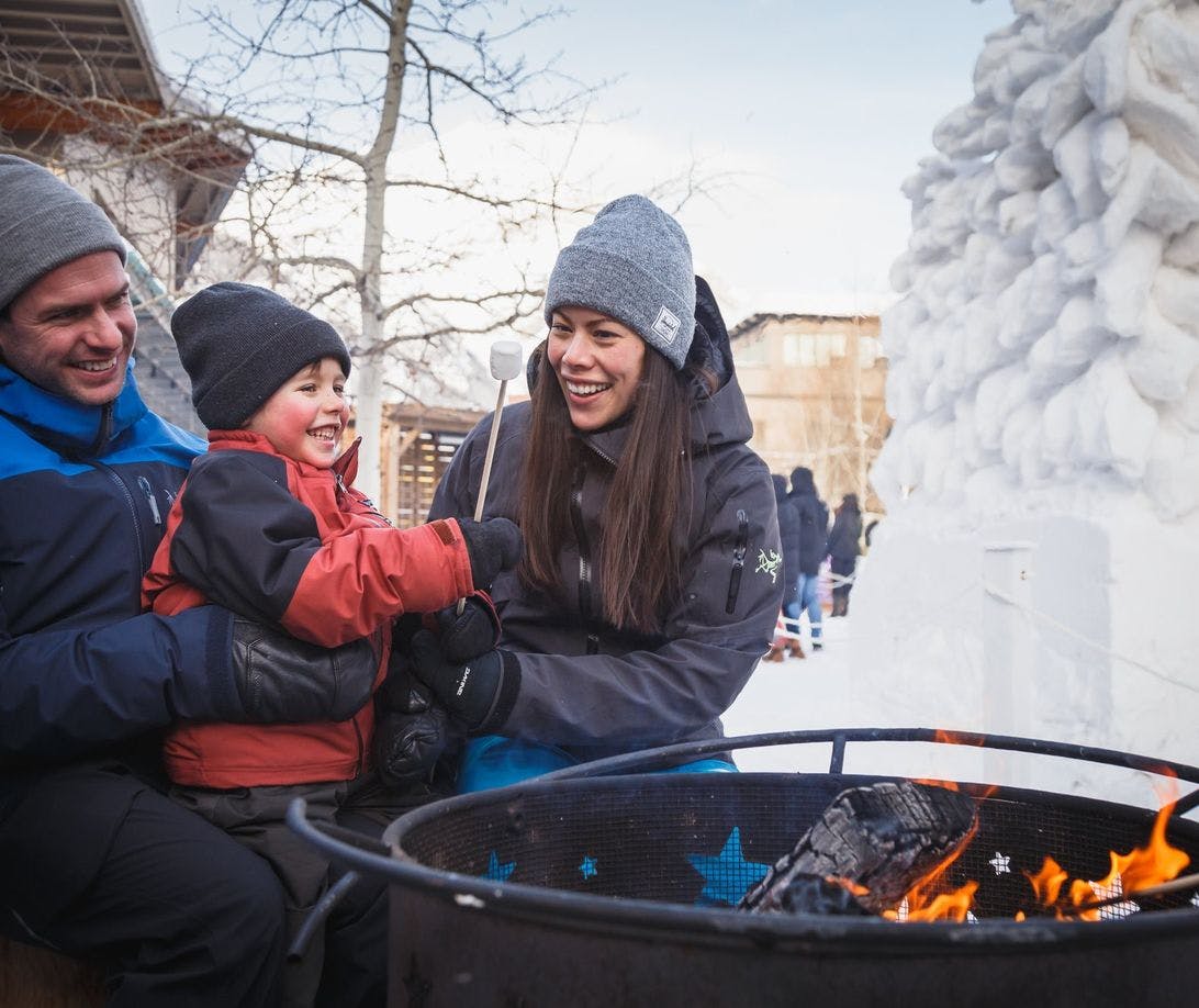 A family wearing lots of cozy winter layers roasting a marshmallow at an outdoor fire pit on a snowy street