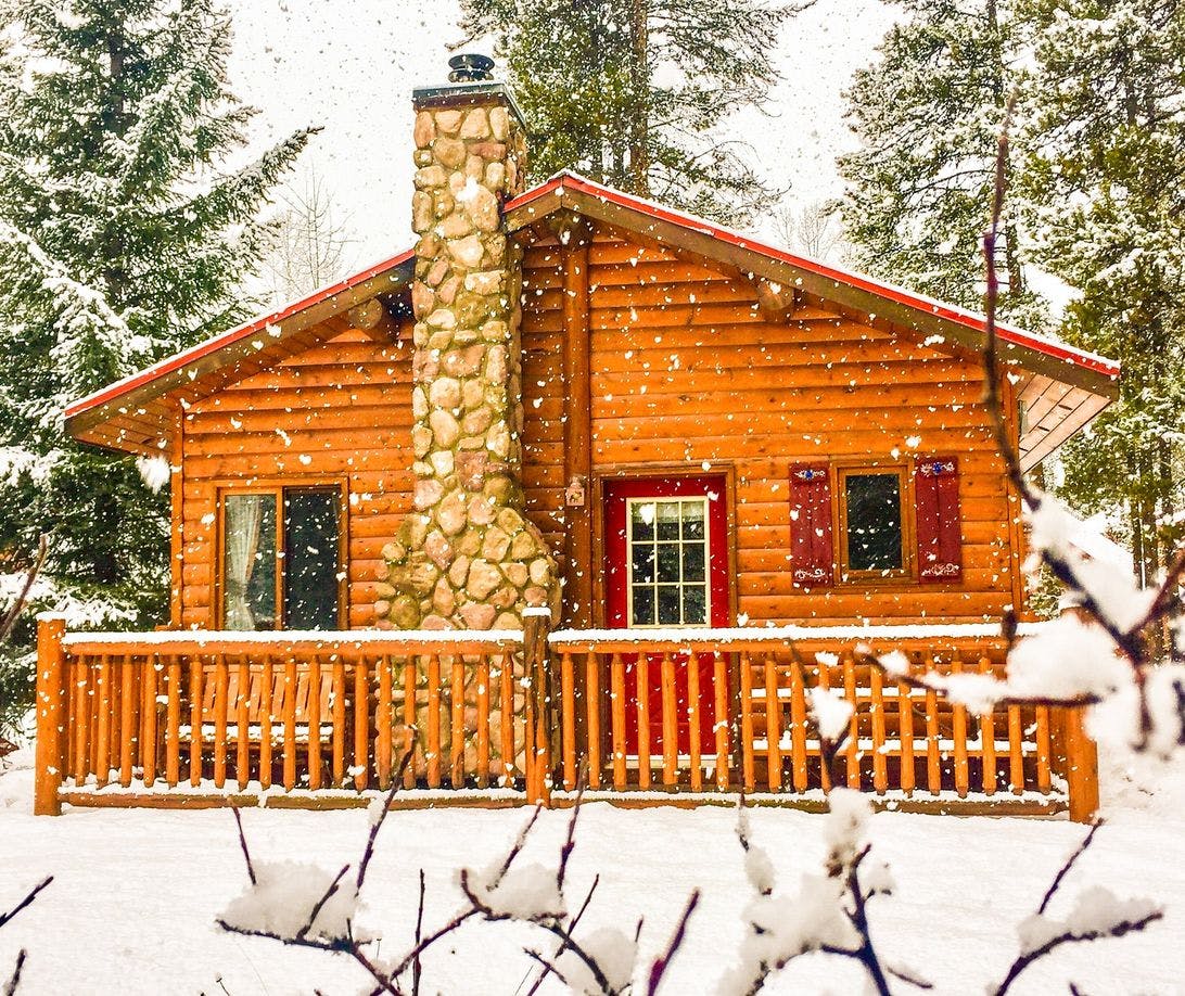 A wooden cabin with a stone fireplace on a snow covered ground as snow continues to fall around it