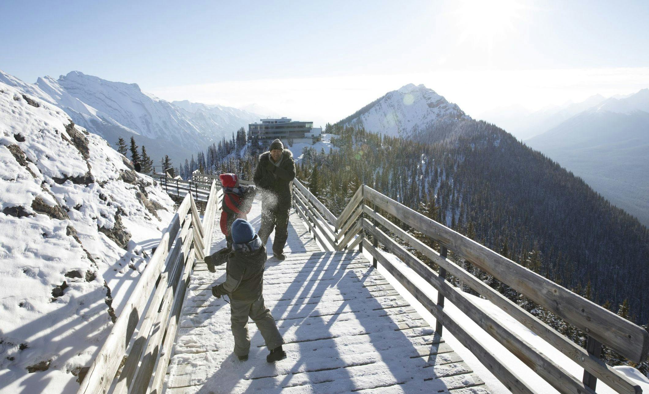 Family playing on the boardwalk at the top of Sulphur Mountain in the winter. Banff Gondola station in the background