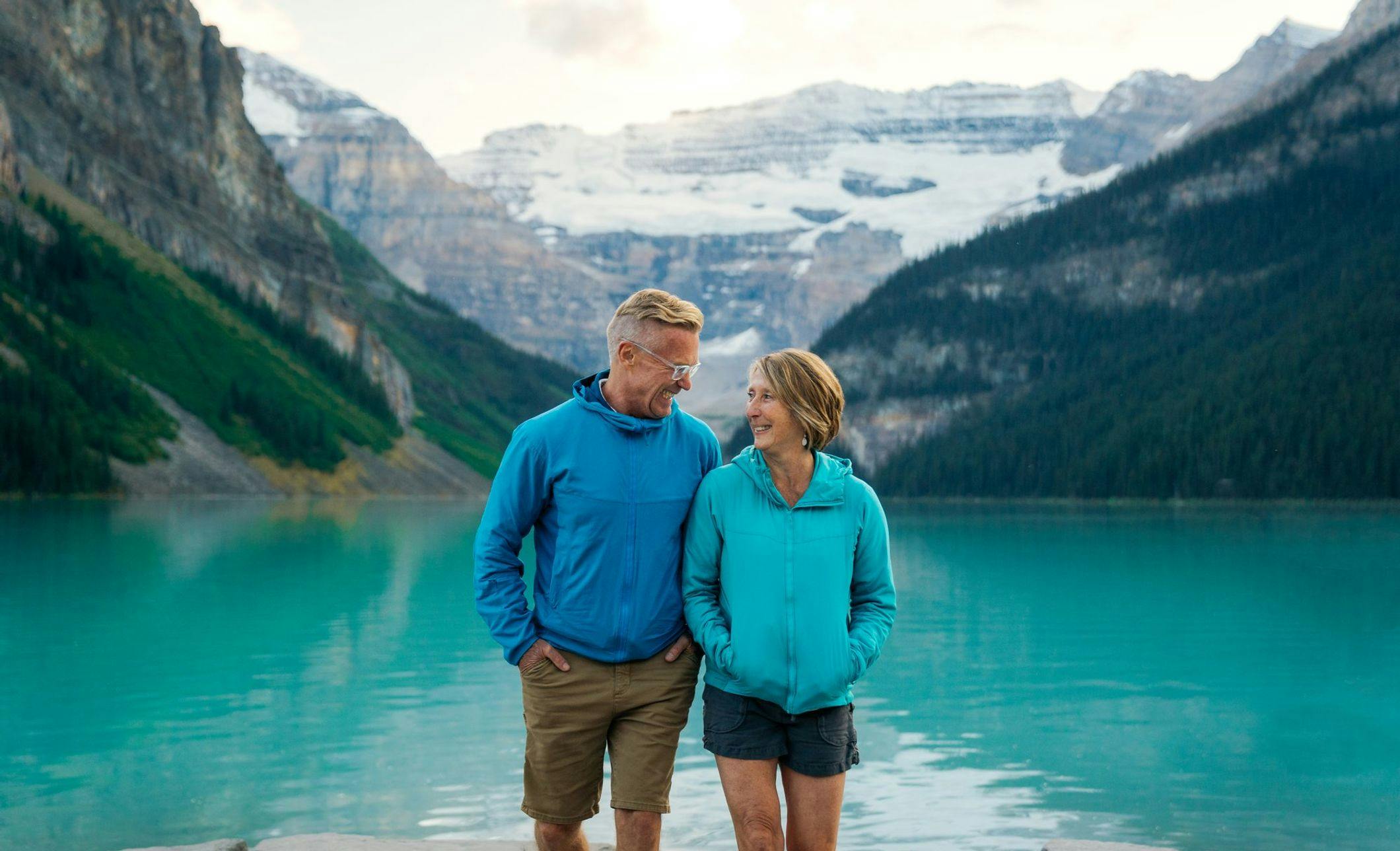 A couple smile and laugh on the shore of Lake Louise with vibrant turquoise water and snow capped mountains (and a glacier) behind them