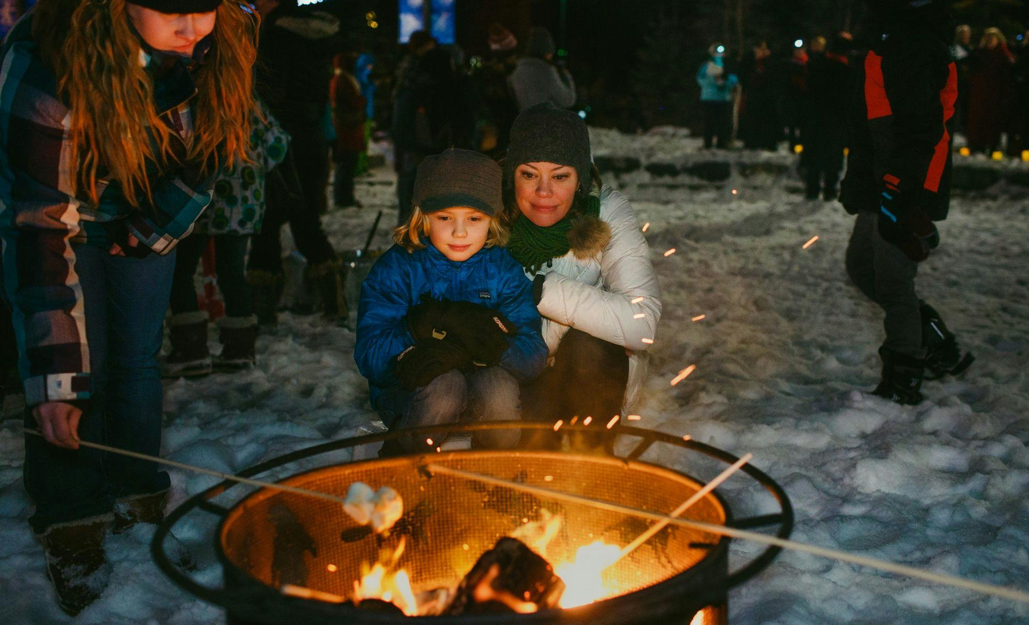 A mom and son sit in front of an outdoor fire pit roasting marshmallows with Christmas lights and snow in the background
