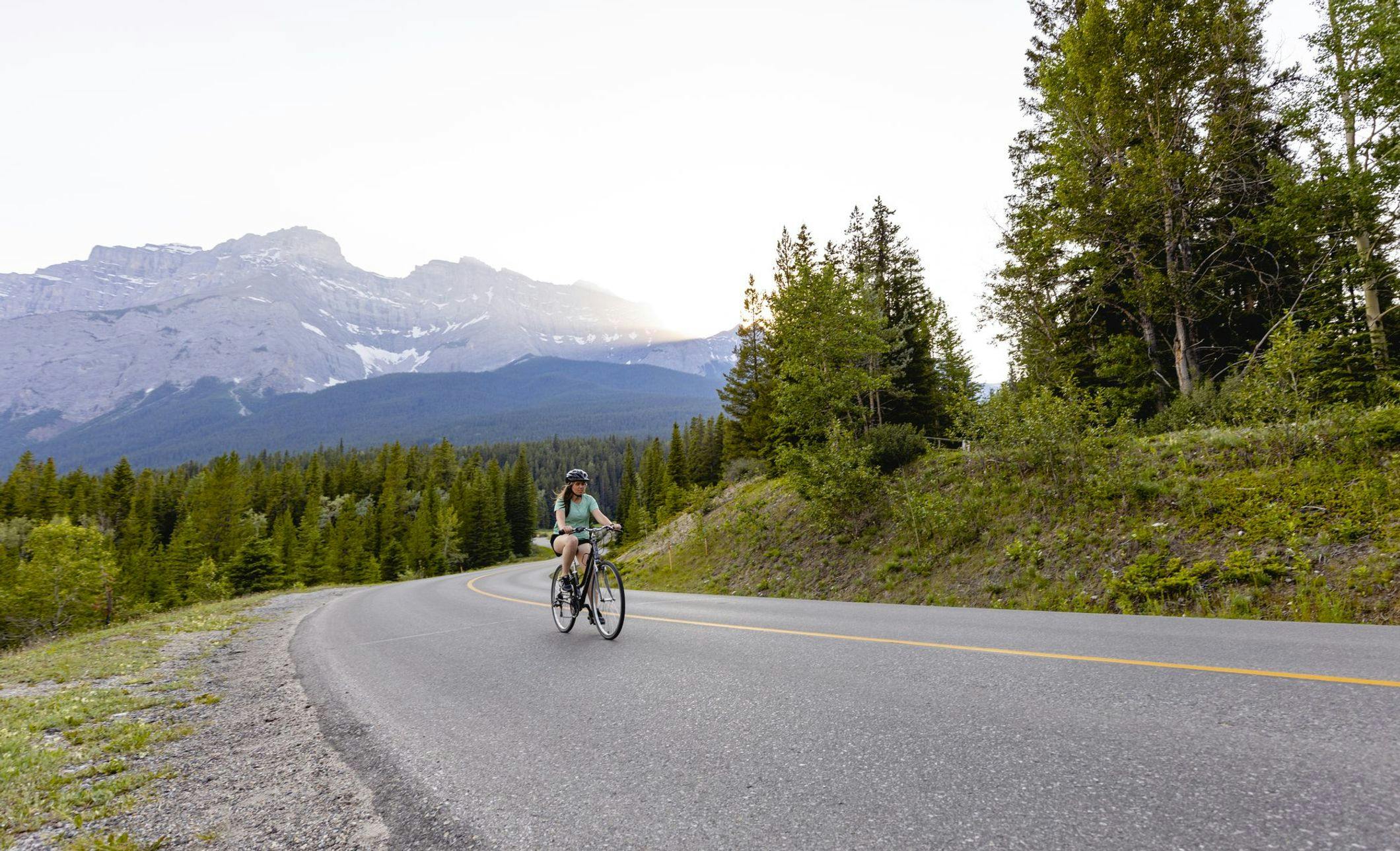 A female rides her bike along an empty stretch of winding road surrounded by mountains on a warm summer day