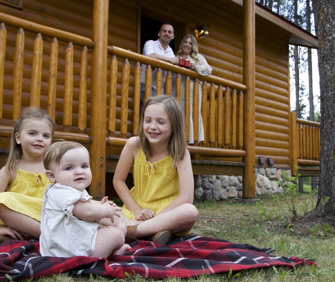 Three young girls on a picnic blanket and parents on the cabin porch behind them