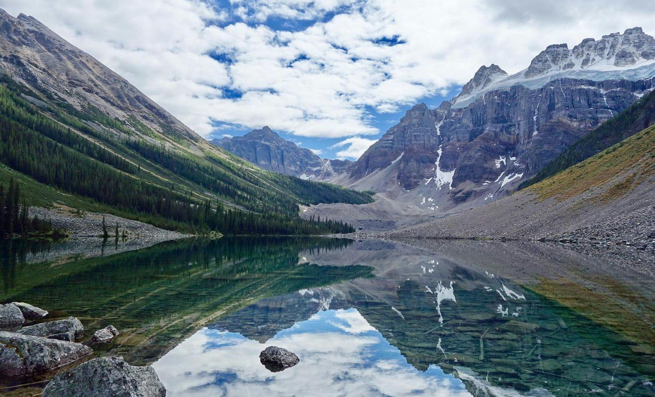 A photo of a clear lake reflecting the trees, snow-capped mountains, and rock formations surrounding it