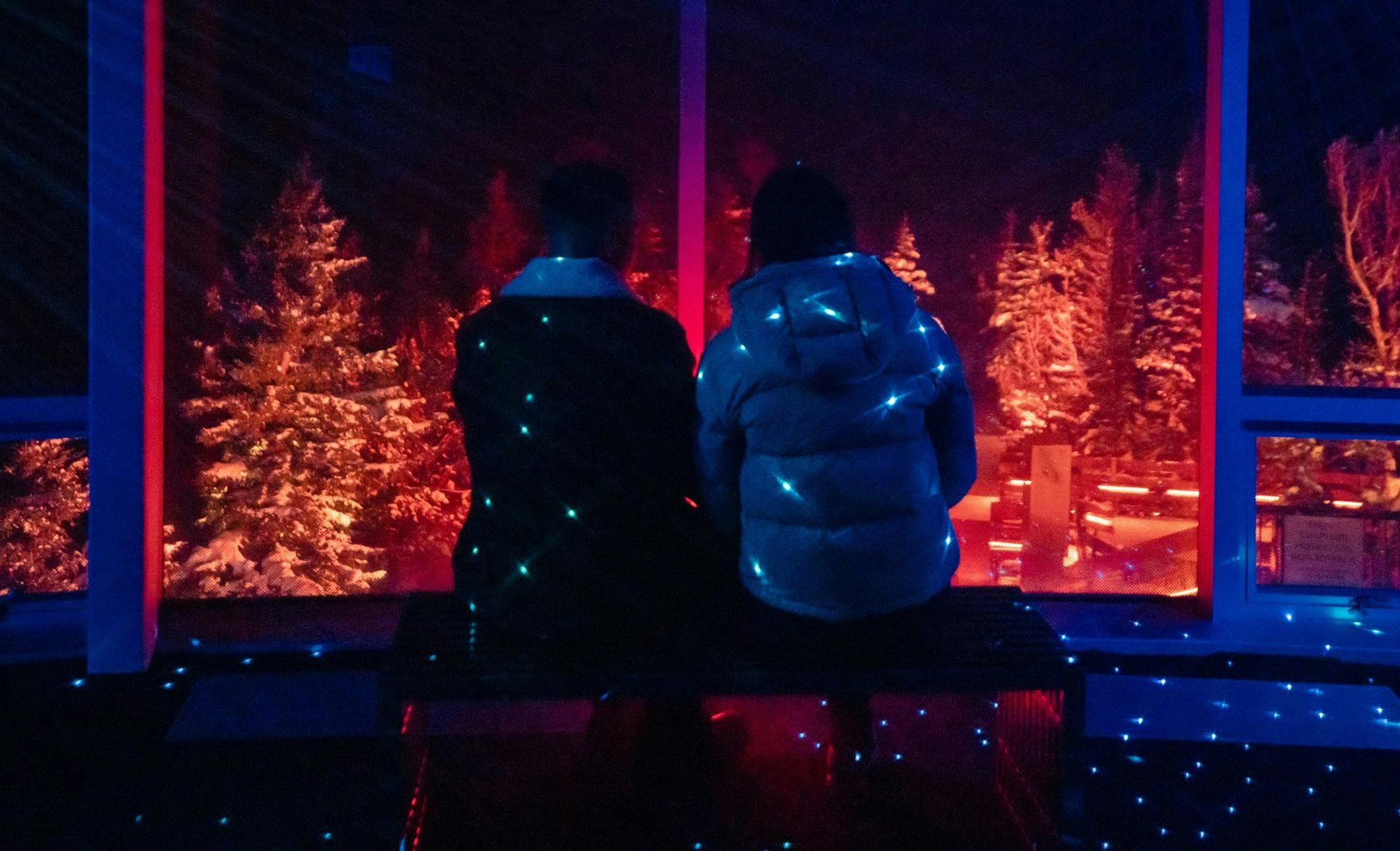 A couple sits on a bench overlooking trees and mountains with starry lights surrounding them
