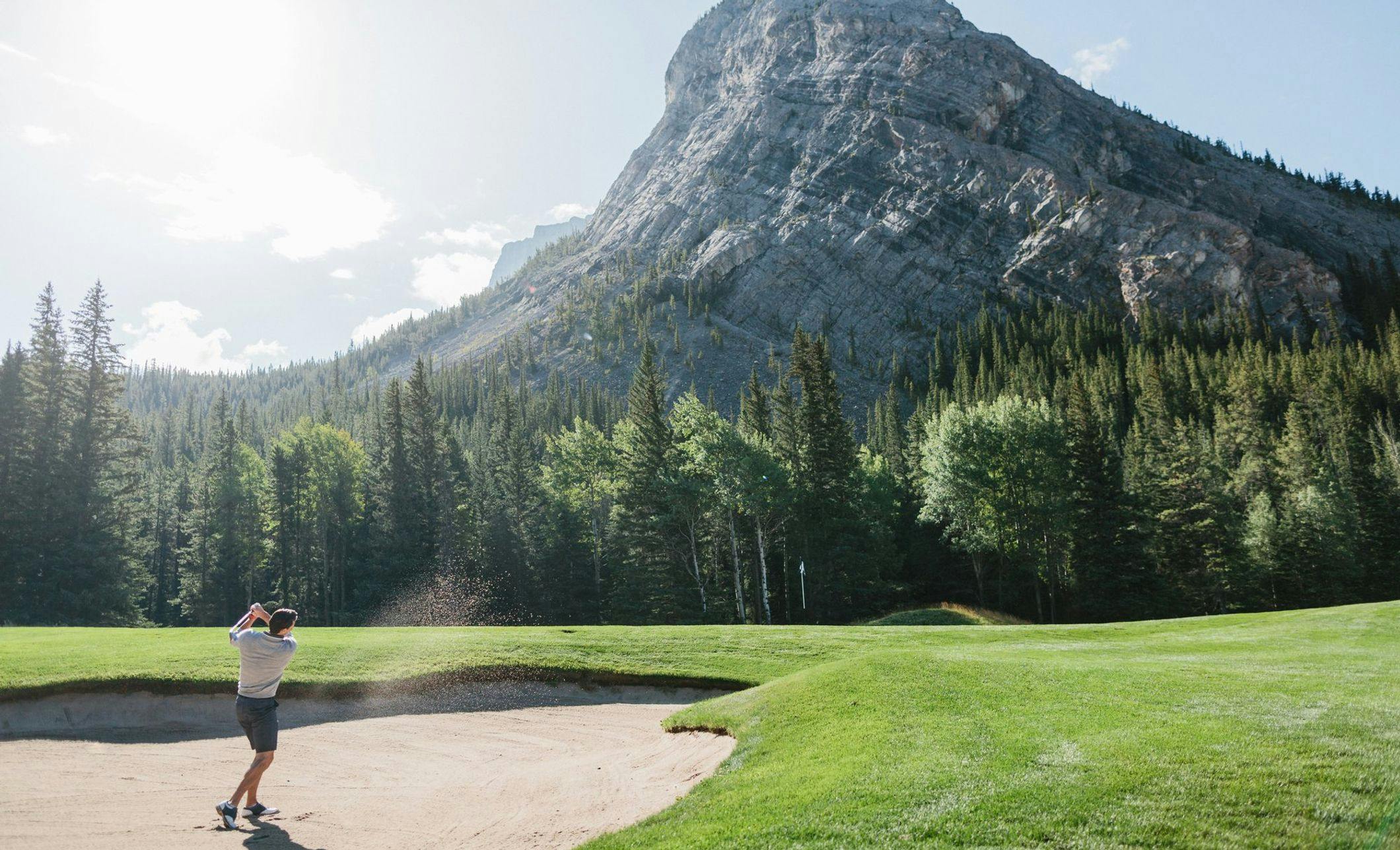 A golfer takes aim on a pristine green course framed by stunning mountains and a clear blue sky
