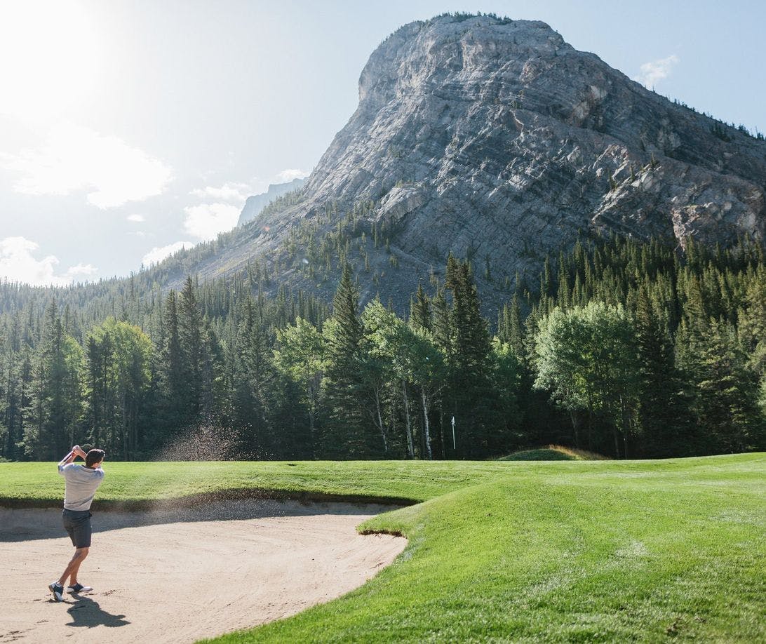 A golfer takes aim on a pristine green course framed by stunning mountains and a clear blue sky