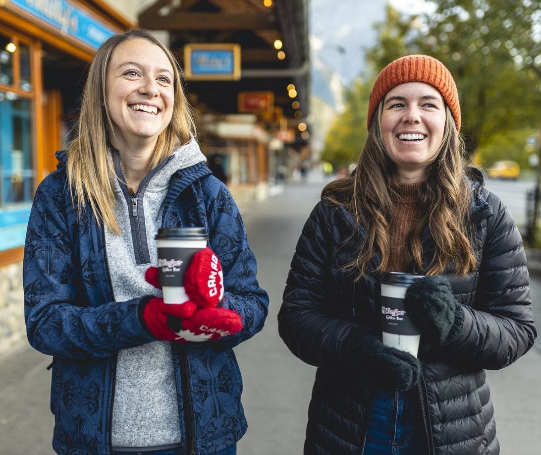 Two friends walking down Banff Ave wearing cozy mittens and holding coffee cups