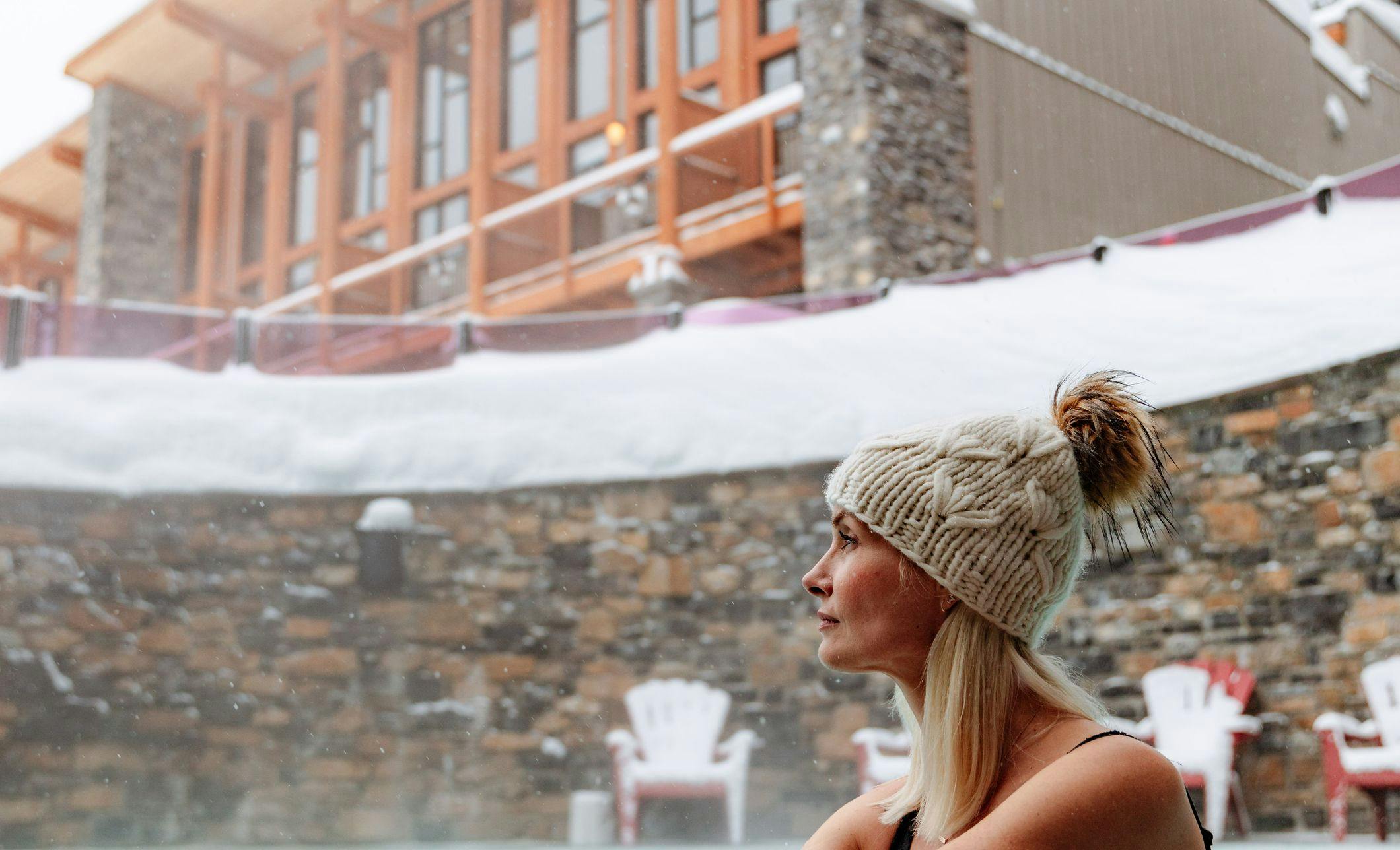 A woman in a toque sitting in an outdoor hot tub in the winter with a ski lodge behind her