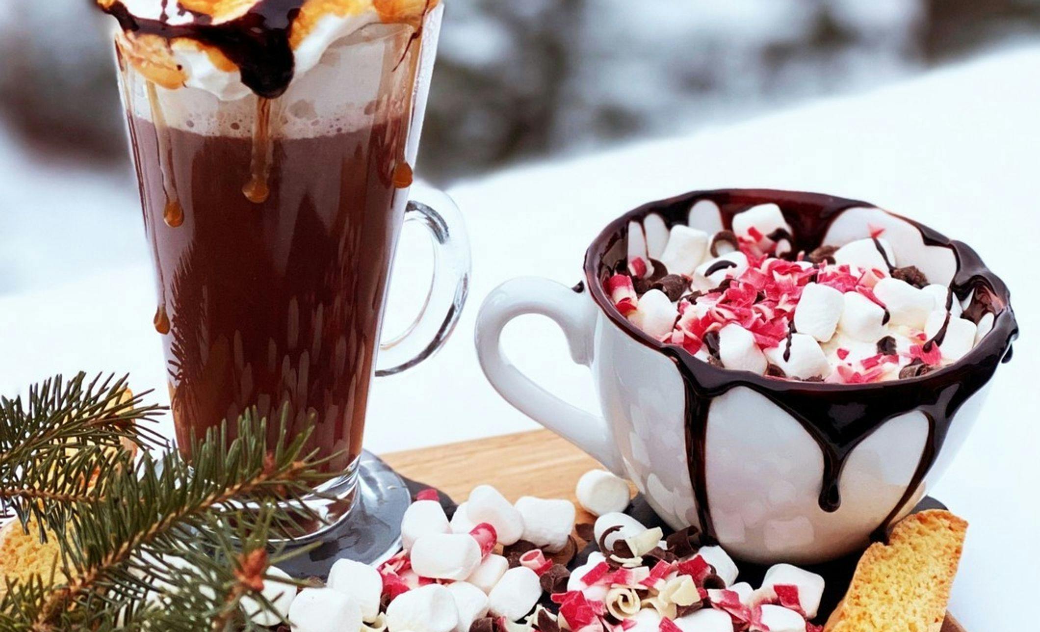 Two hot chocolates set outside in the winter with festive decorations and toppings
