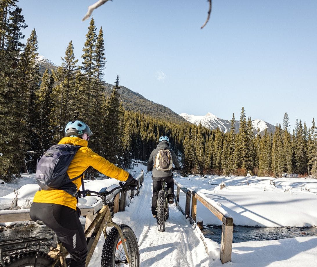 Two cyclists on fat bikes traveling across a snow covered bridge