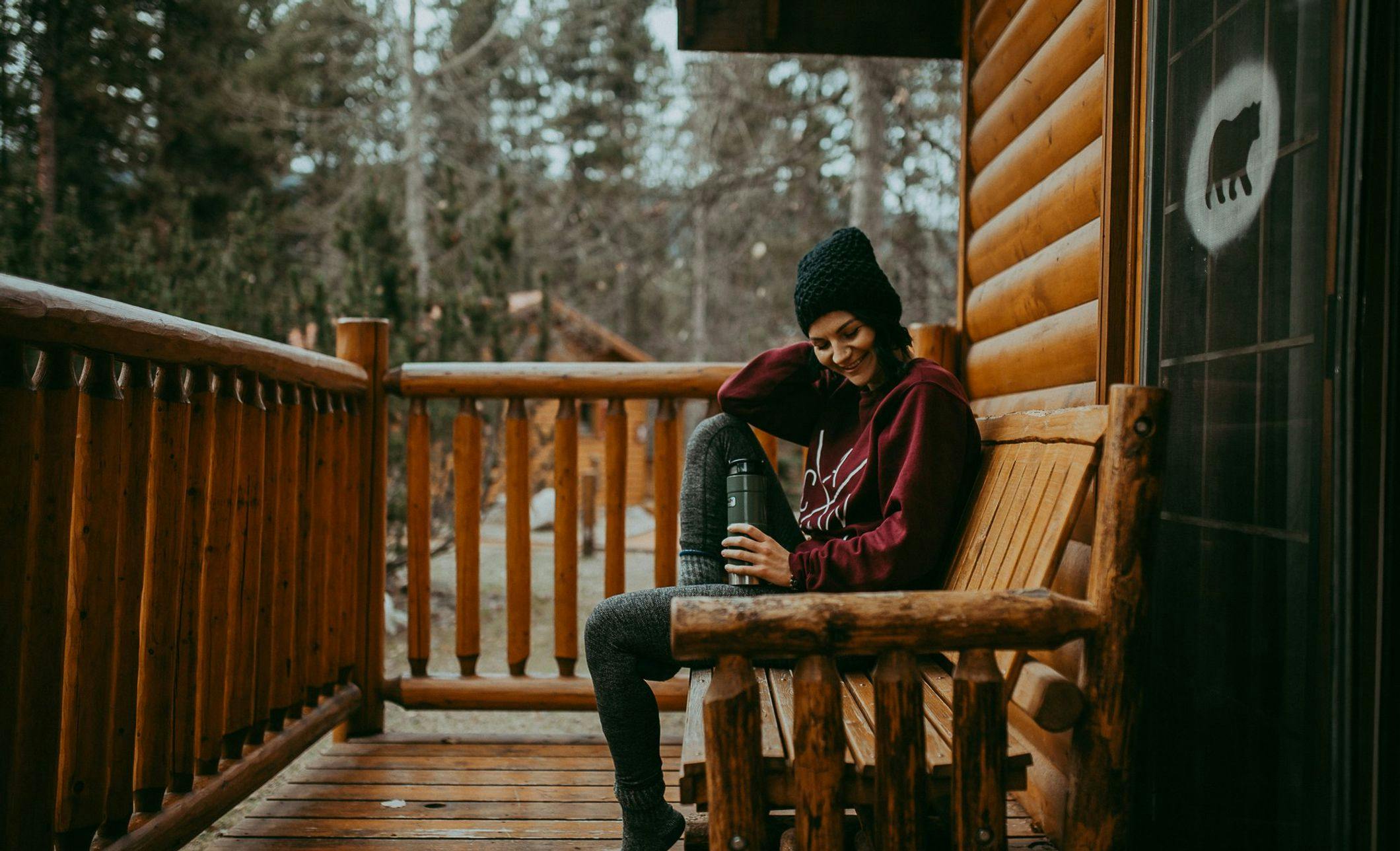 Woman sitting outside a cozy, wooden cabin wearing sweats, a toque, and drinking coffee