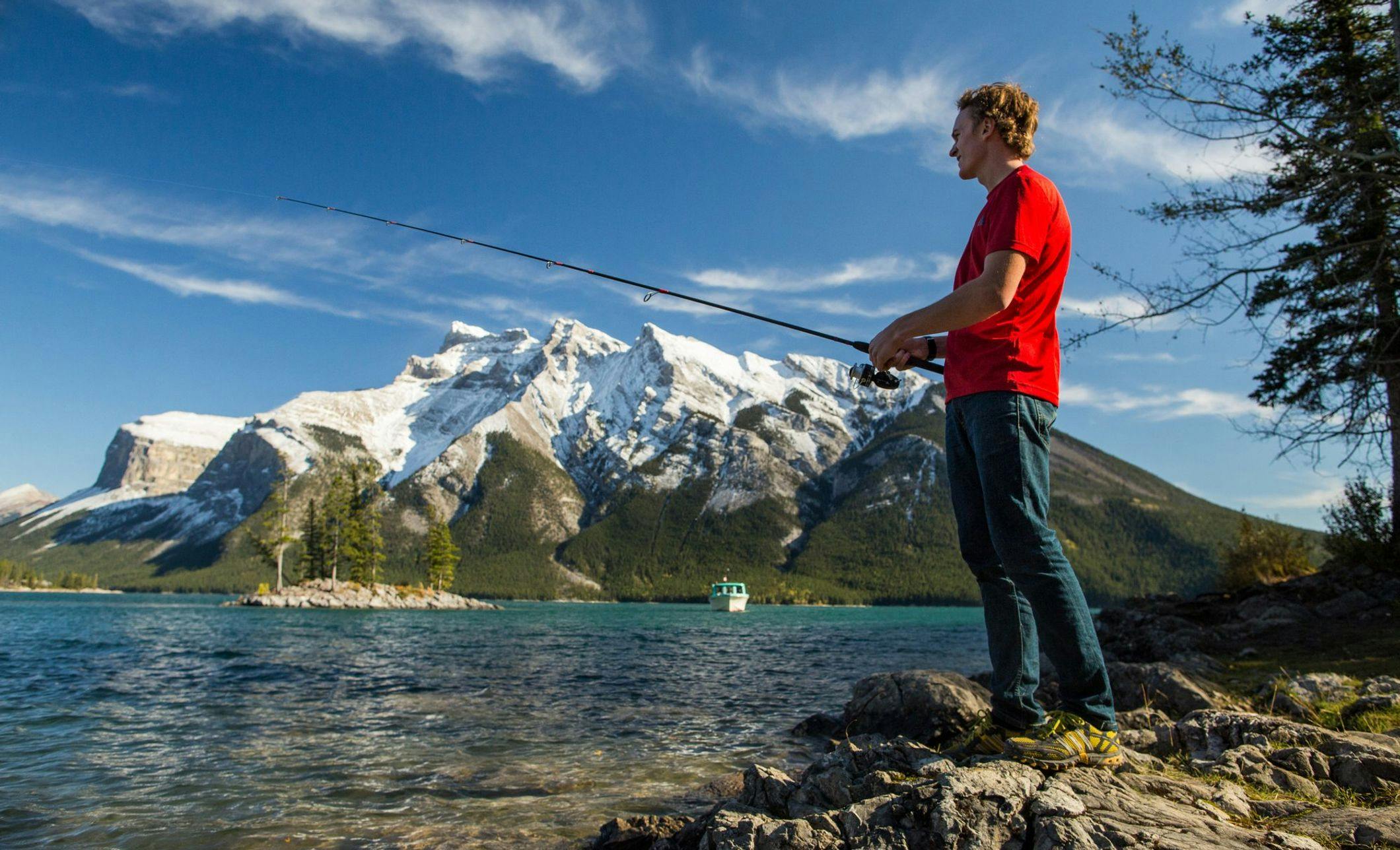 A man in tan pants and a red t-shirt fishes in a stunning blue lake framed by mountains behind him