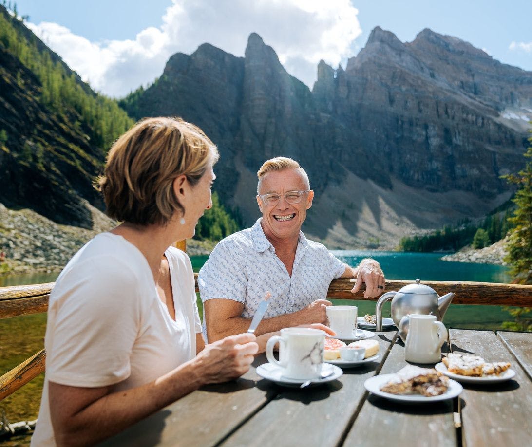 A mature couple sits at a picnic table enjoying tea and snacks overlooking a turquoise lake framed by mountains