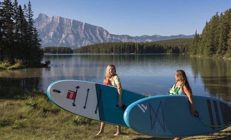 Two friends walk with paddleboards in hand to a stunning turquoise lake surrounded by mountains