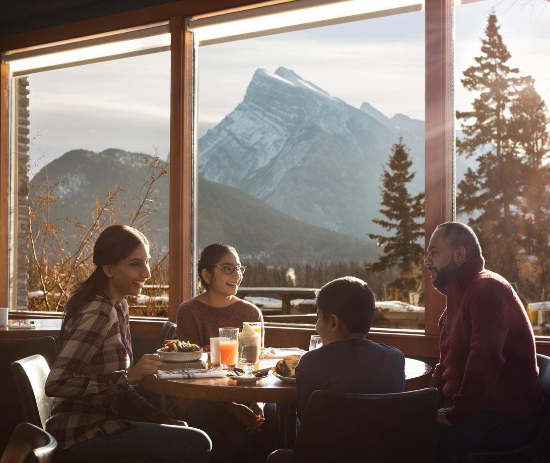 A family of four sit around a table laughing and enjoying brunch with views of a large mountain outside the window on a sunny day