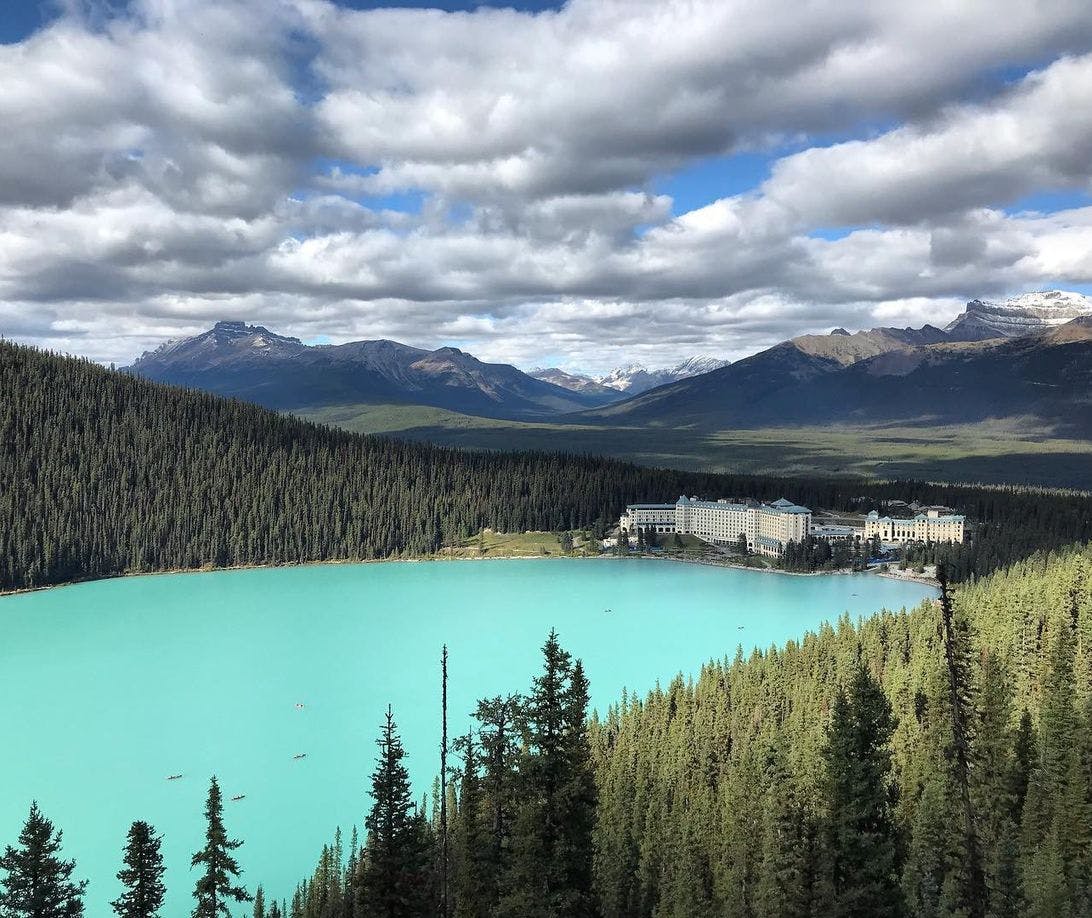 Photo taken from a viewpoint overlooking a vast turquoise lake (Lake Louise) with a large hotel (Chateau Lake Louise) on the edge in front of a large mountain range