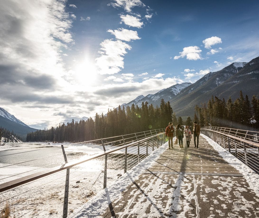 A group of friends walk across a bridge with snow capped mountains surrounding them 