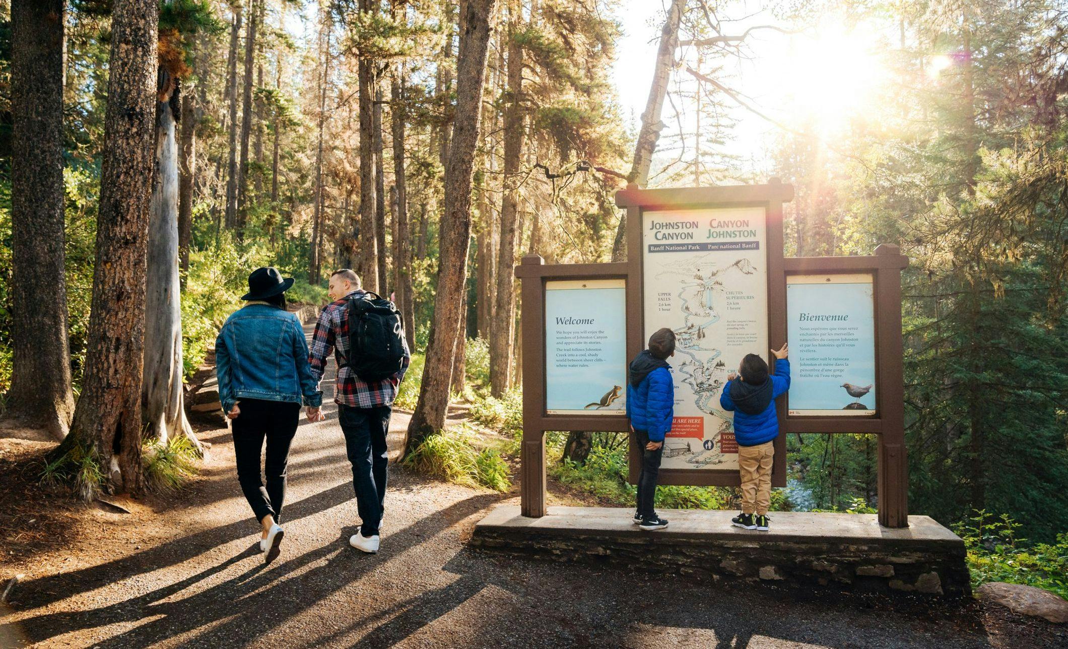 A couple walks along a trail while their two young boys stop to read the sign titled &quot;Johnston Canyon&quot;