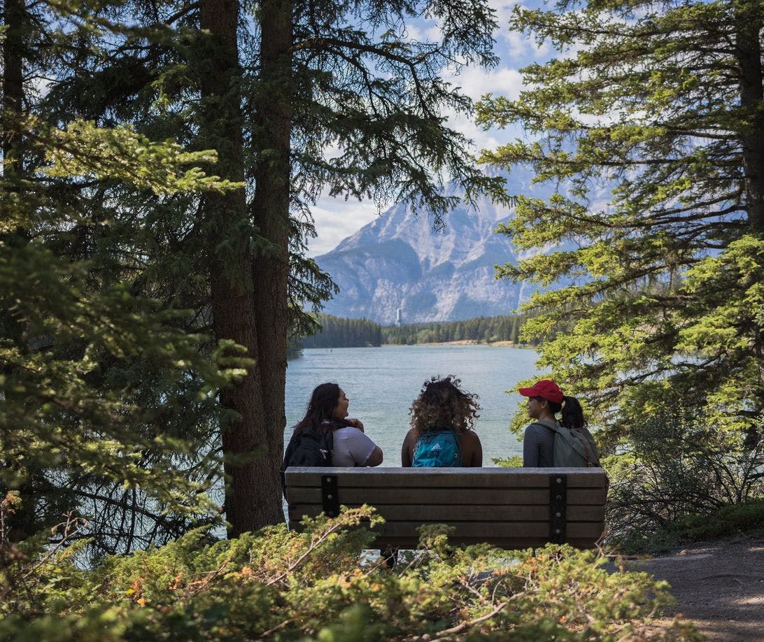 Three friends sit on a bench in a forested area looking out at a glacial blue lake