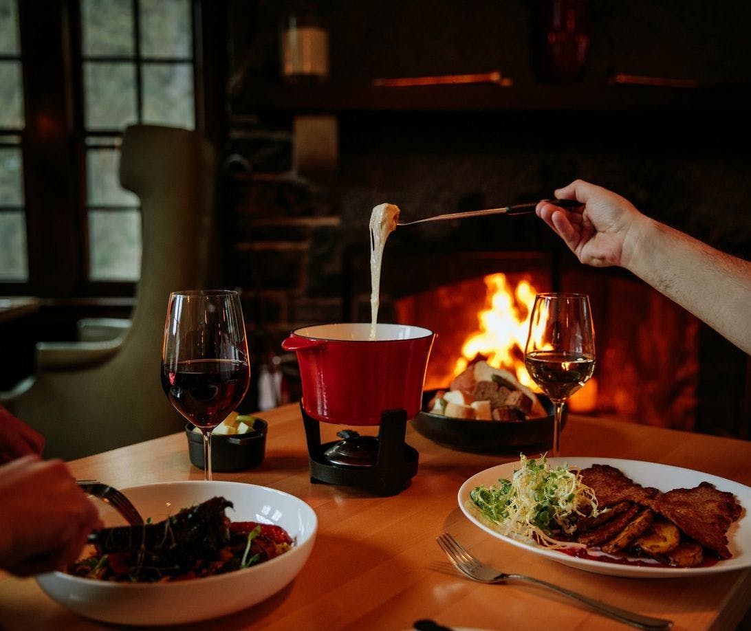 A couple sits at a table enjoying appetizers and a large pot of cheese fondue with a fire crackling in the background