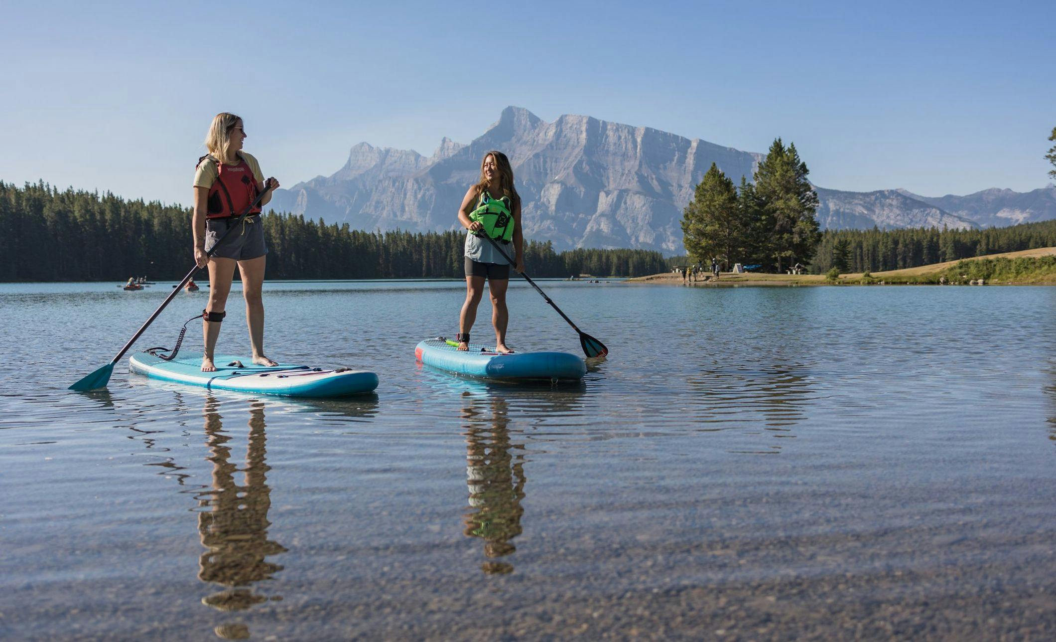 Two female paddleboarders balance on their boards on a clear blue lake with people enjoying the lake front behind them