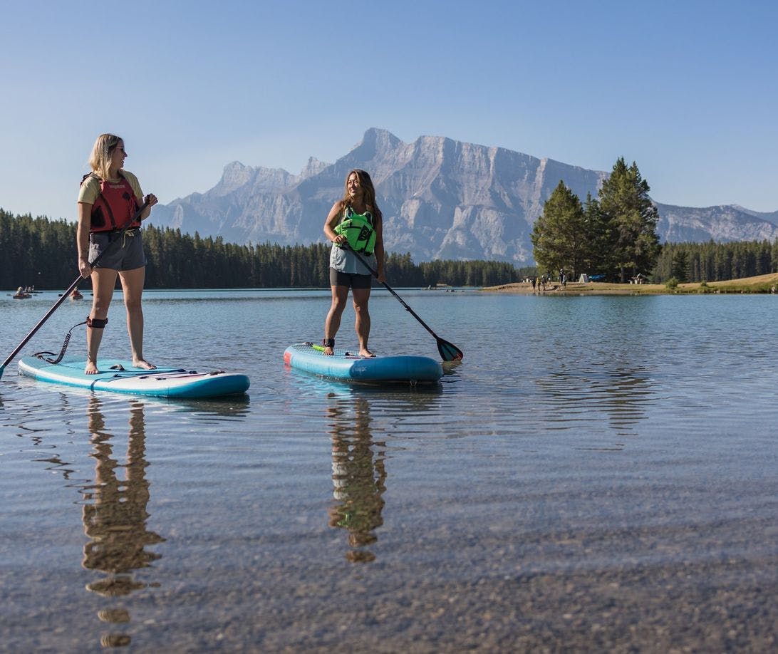 Two female paddleboarders balance on their boards on a clear blue lake with people enjoying the lake front behind them