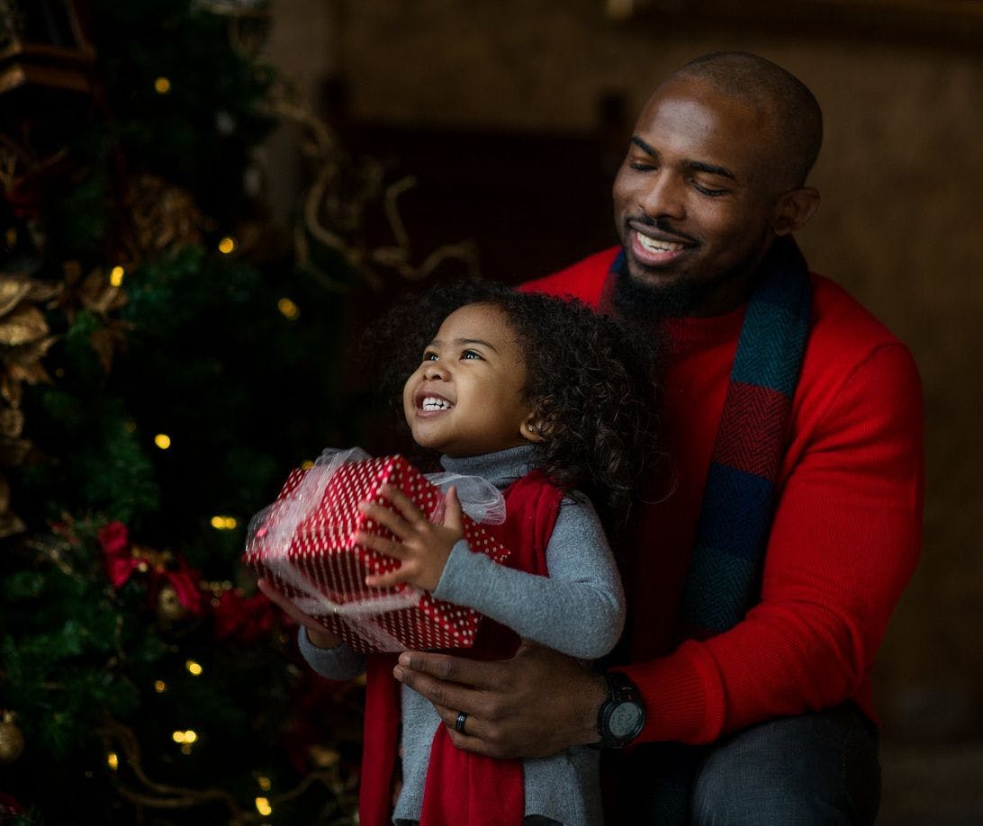 A father and daughter smiling and holding a Christmas gift next to a decorated Christmas tree