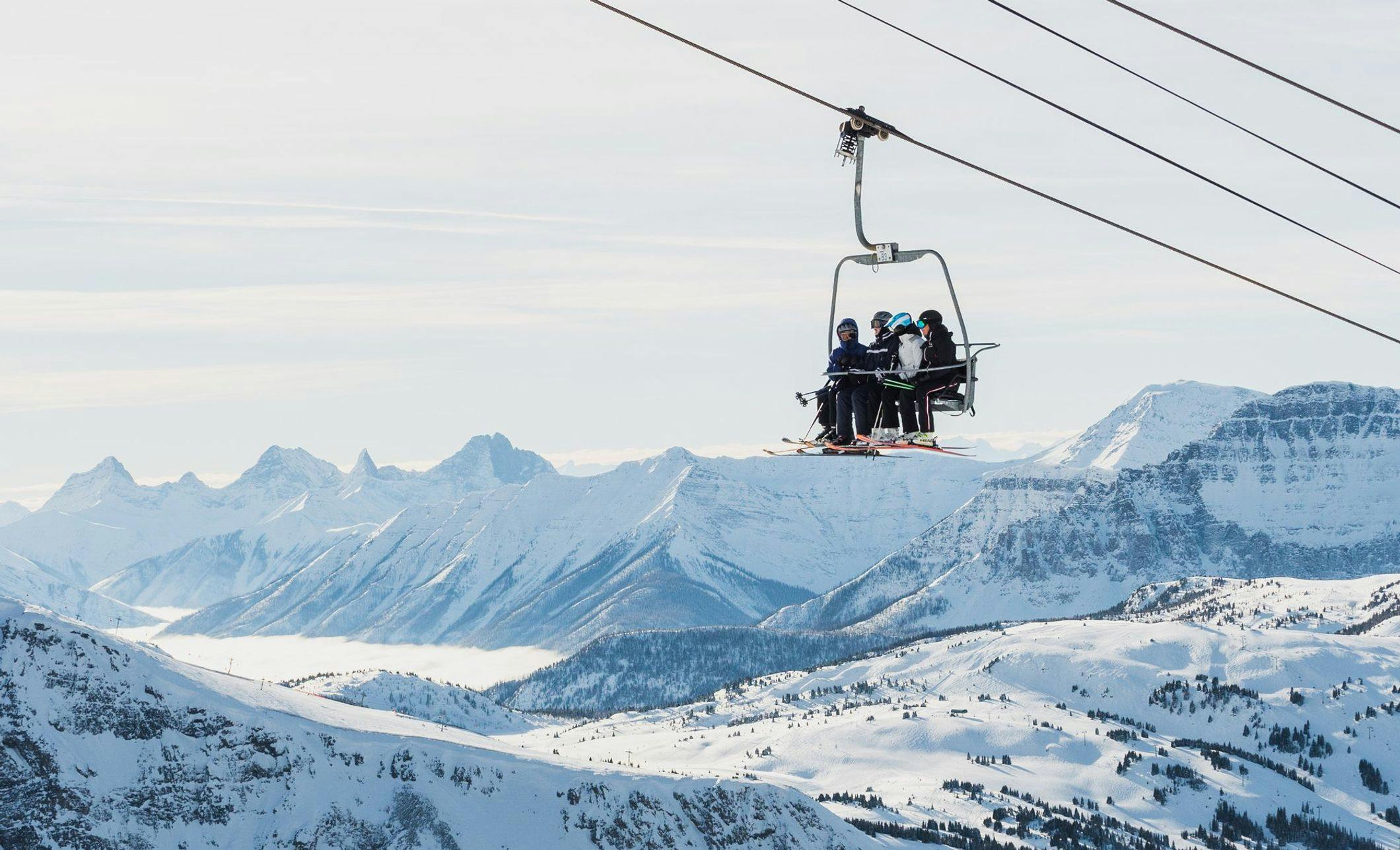 4 friends ride an open chairlift over a snowy ski hill with mountains in the background