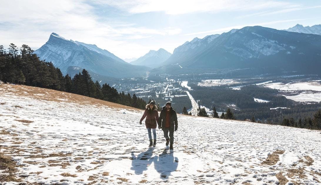 A couple hiking on a hill with grass peeking through the snow and an aerial view of the town and the mountains behind them
