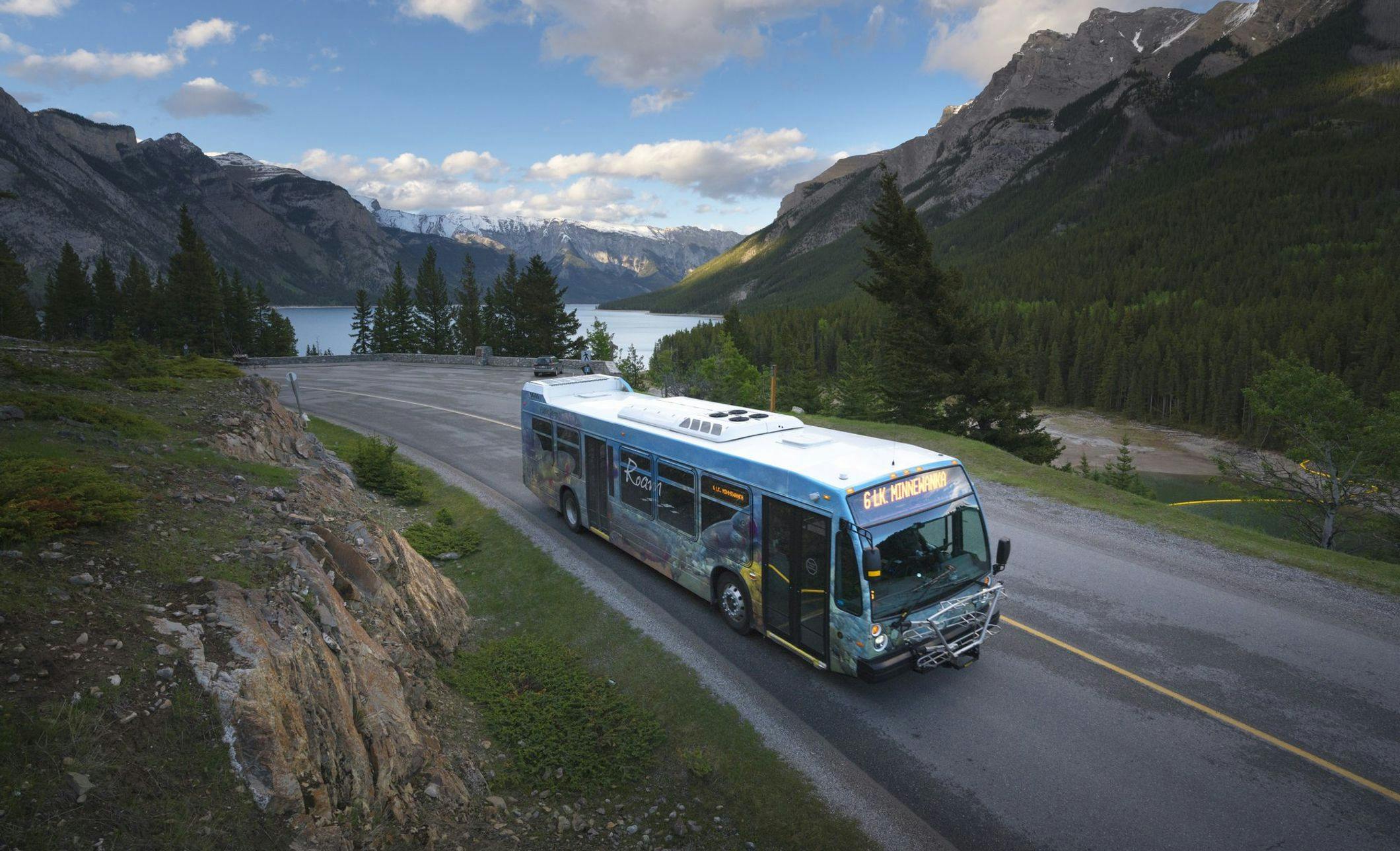 A public bus travels along a road with mountains and blue lakes in the background on a sunny summer day