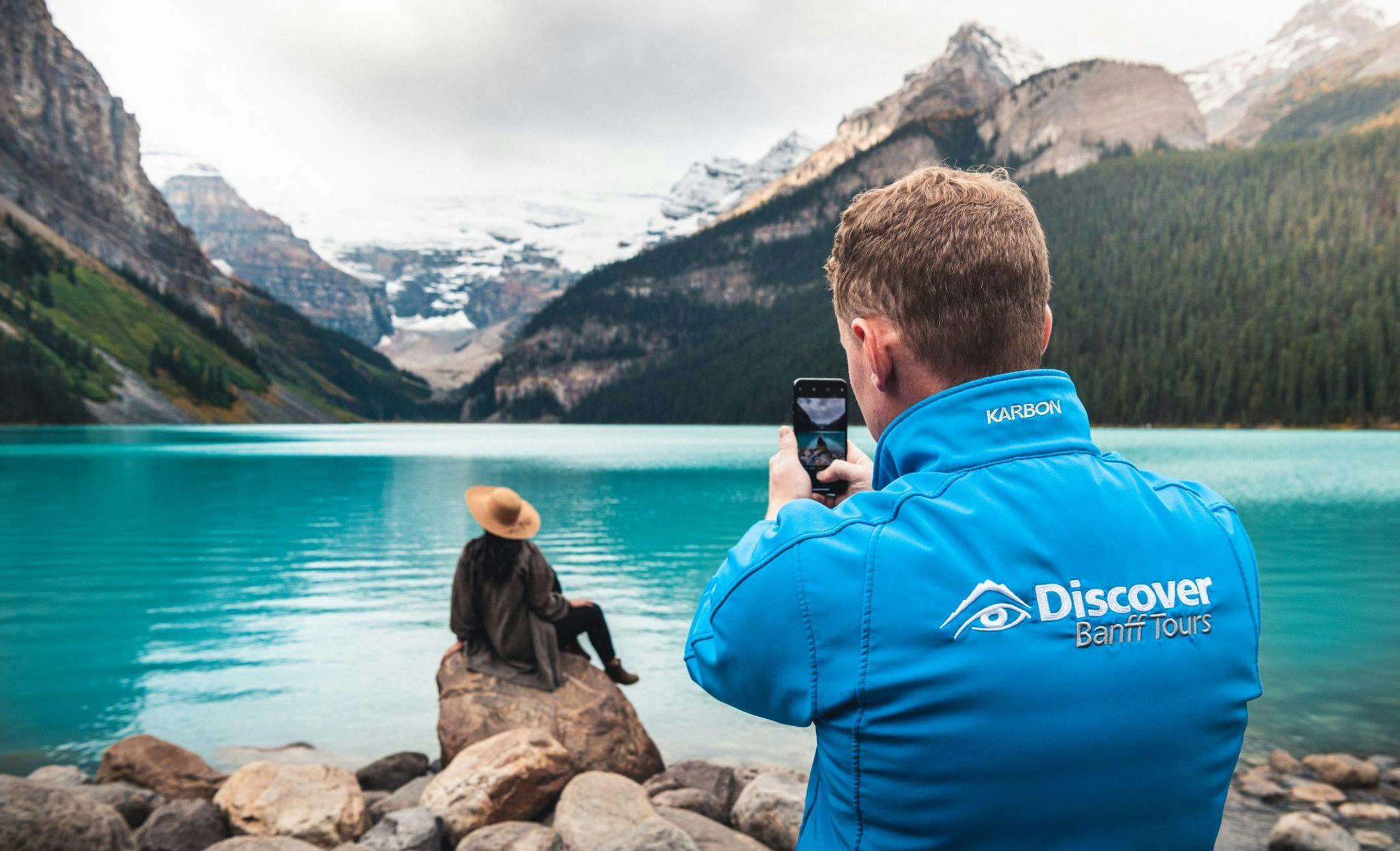 A tour guide takes a photo of a traveler in front of a blue lake and a large glacier as she poses on some rocks at the foot of the lake