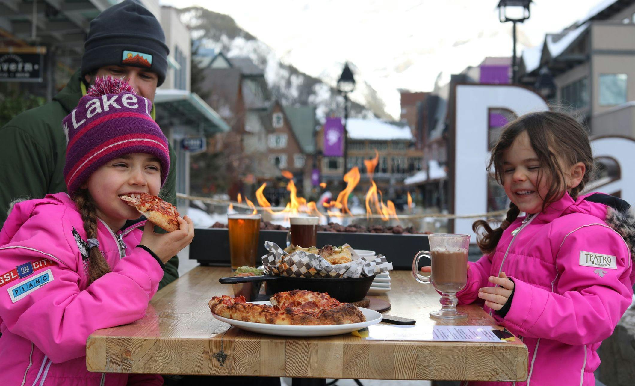 A dad and two young girls enjoy pizza next to an outdoor fireplace 
