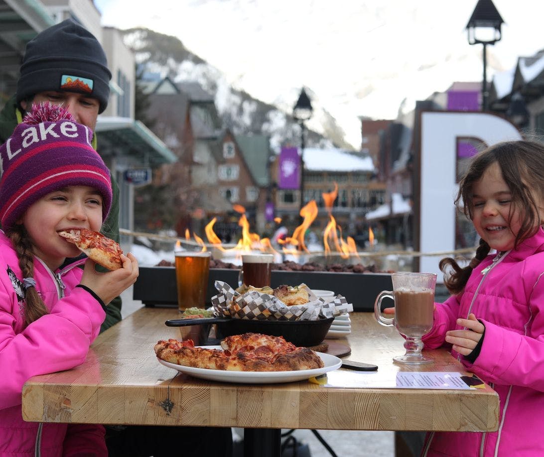 A dad and two young girls enjoy pizza next to an outdoor fireplace 