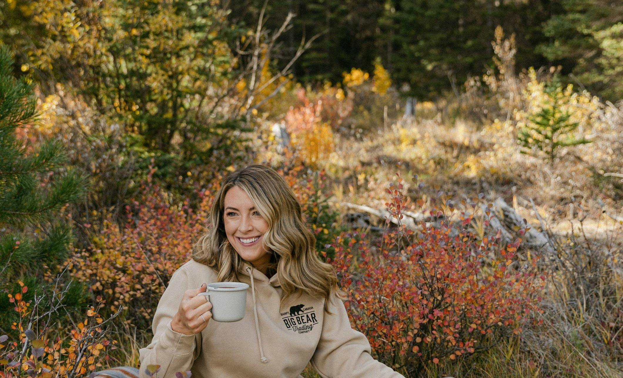 Female relaxing on a cozy blanket with a cup of coffee and an oversized sweater. Fall foliage in the background
