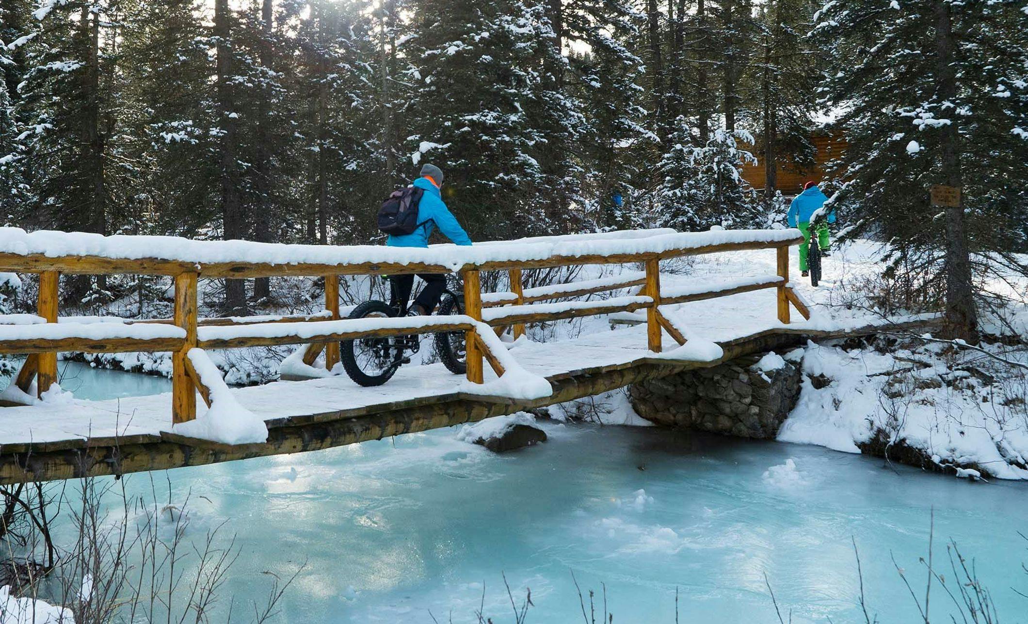 Two cyclists riding fat bikes across a snow covered bridge and over a frozen river