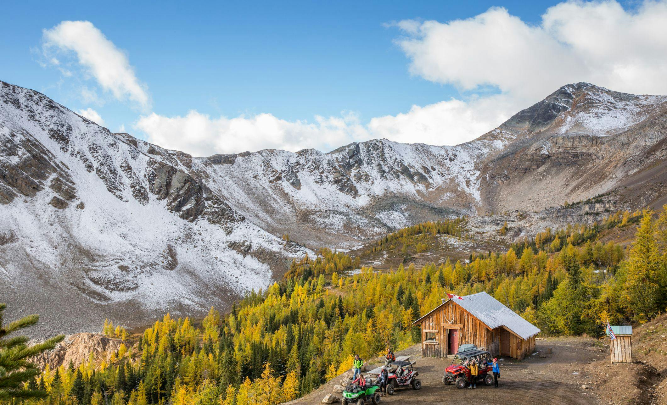 ATVs are parked next to a wooden cabin and surrounded by snow capped mountains and lush yellow and green trees