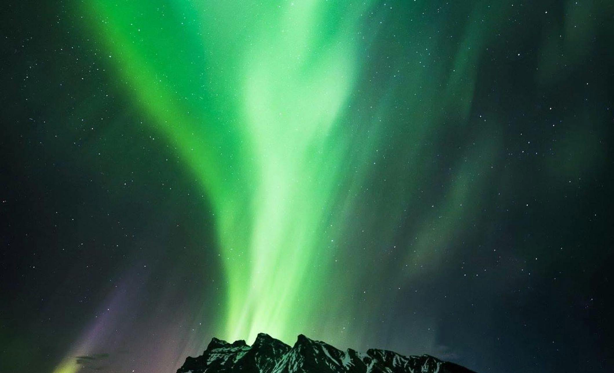 Green northern lights swirl in the sky above a snowcapped mountain overlooking a lake at night