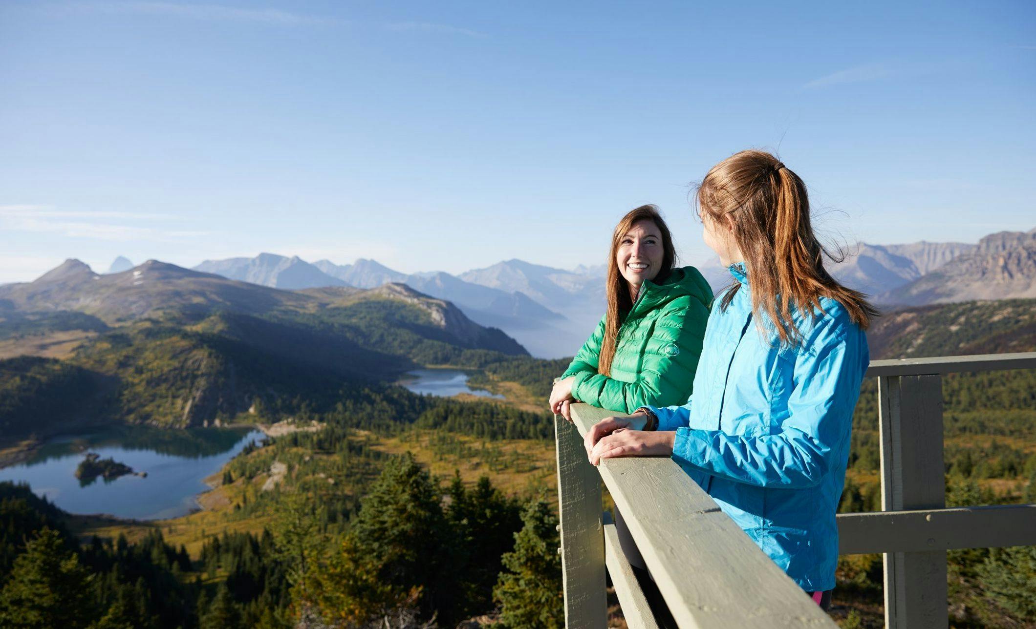 Two friends stand on a lookout deck with alpine lakes and hiking trails surrounding them. They are wearing puffy coats despite the sunshine as they are in the alpine