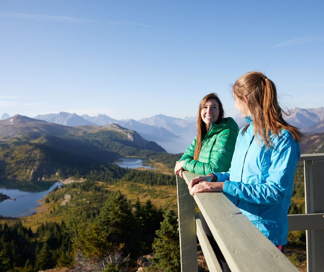 Two friends stand on a lookout deck with alpine lakes and hiking trails surrounding them. They are wearing puffy coats despite the sunshine as they are in the alpine