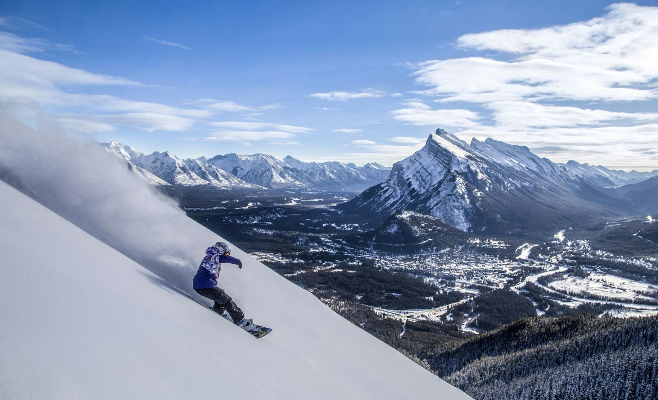 Snowboarder heading down a mountain with a snow covered town of Banff below and snowy mountains surrounding